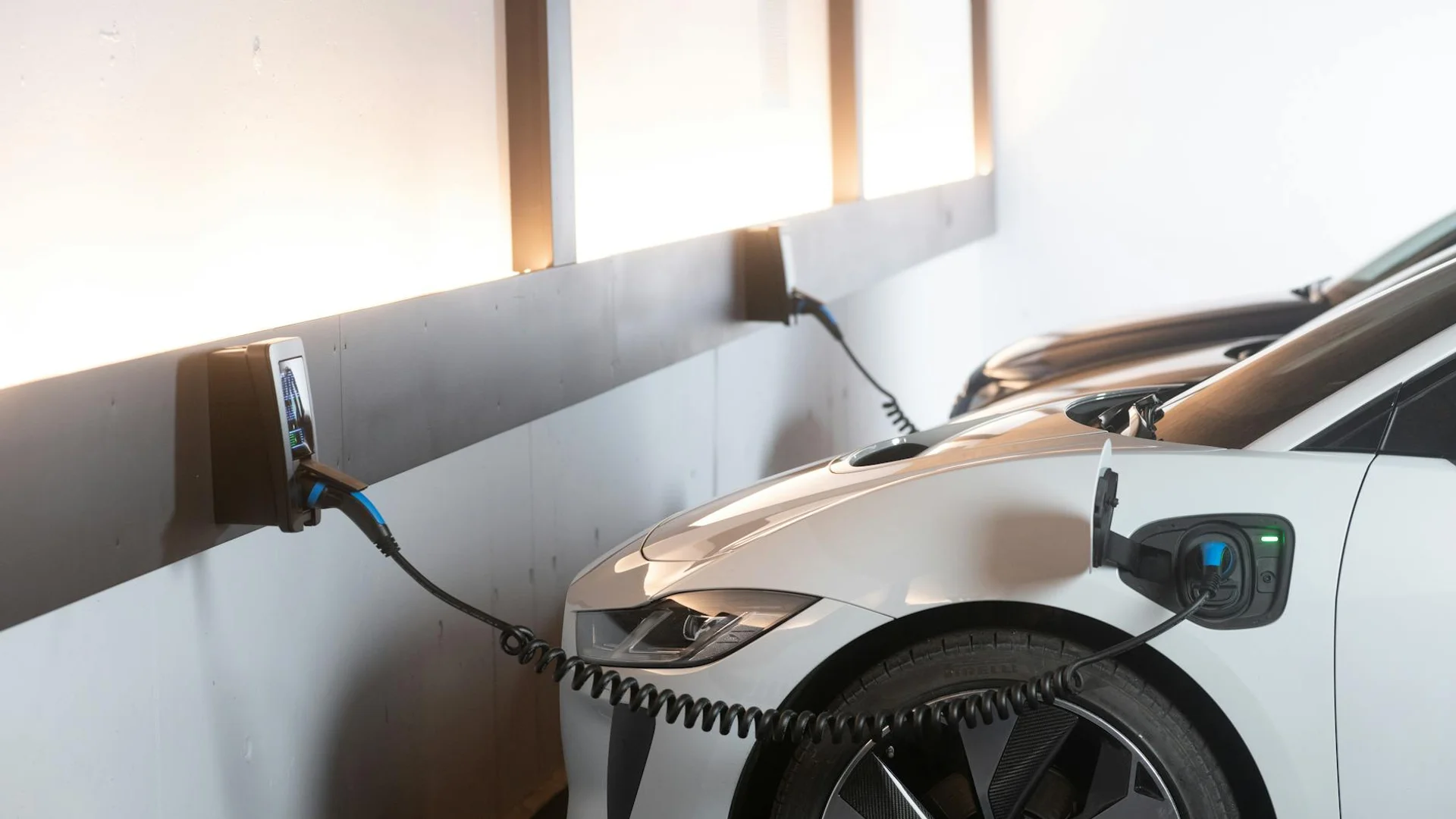 A white electric car is charging from a wall-mounted station with a blue and black cable.
