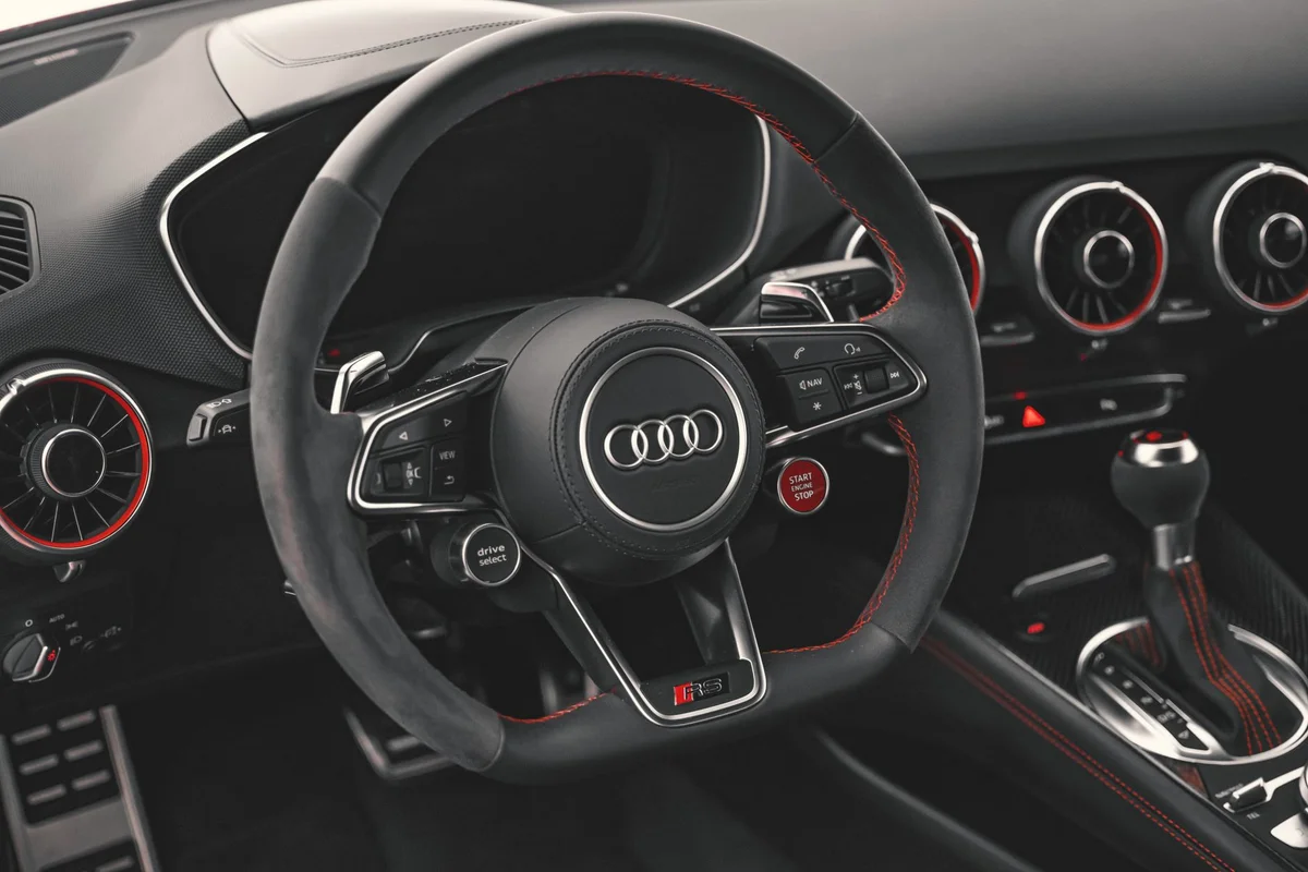 A black car interior shows a steering wheel with red stitching, an Audi logo, and a red start/stop button.