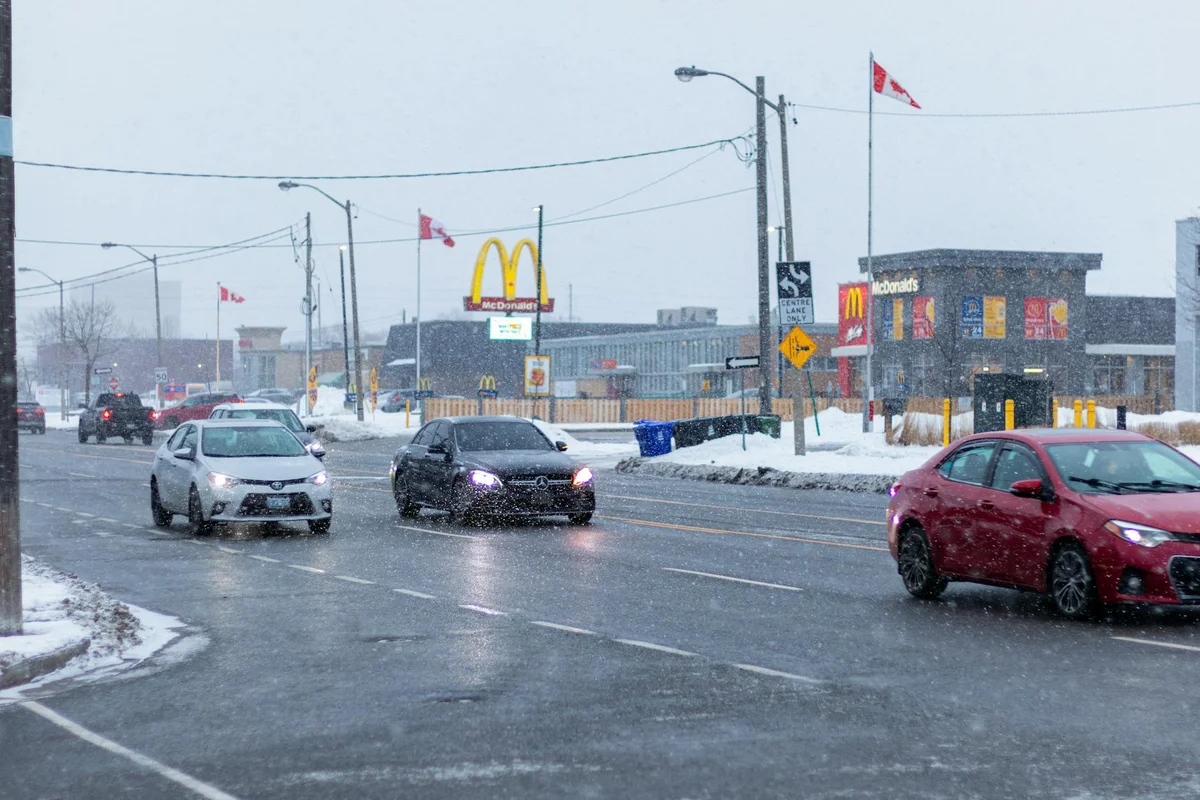 Cars drive on a wet road with snow falling, a McDonald's building and sign are visible in the background.