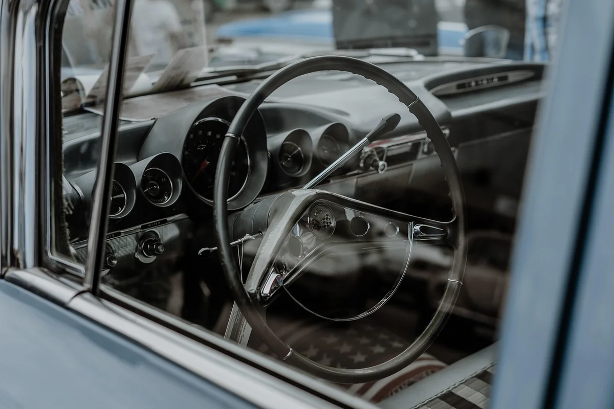 The interior of a vintage car is visible through the driver's side window, showing a black steering wheel and dashboard.