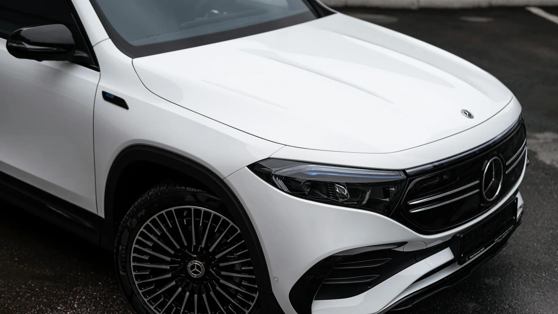 A white Mercedes-Benz car is parked on a wet asphalt surface, showing its front right side and headlight.