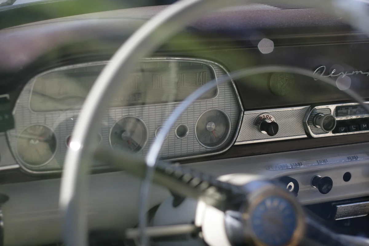 The dashboard of a vintage car, with a white steering wheel in the foreground and a textured silver panel.