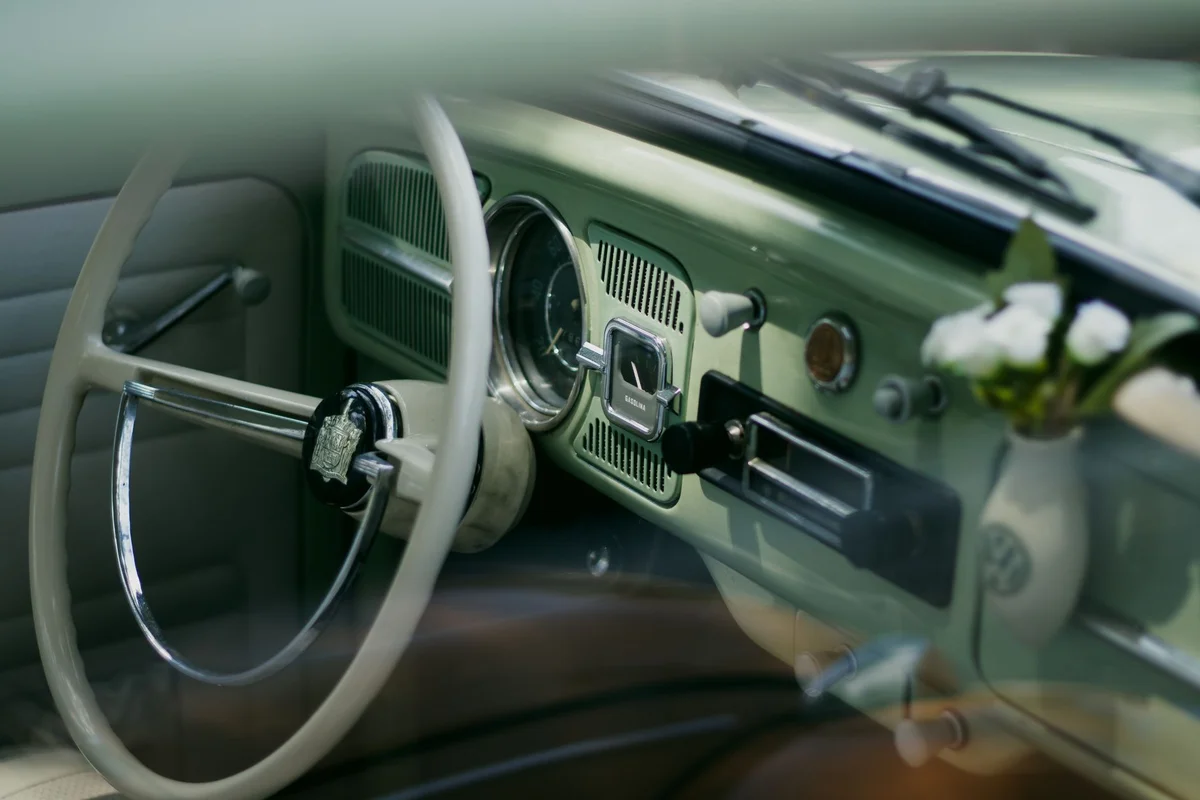 A light green vintage car interior with a cream steering wheel and a small vase of white flowers on the dashboard.
