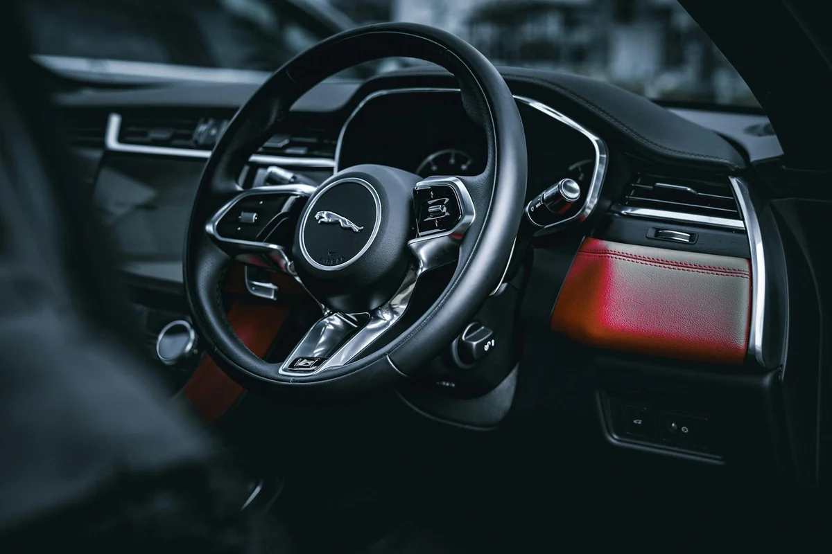 A black car steering wheel with a silver Jaguar logo is centered, with a red leather dashboard panel on the right.