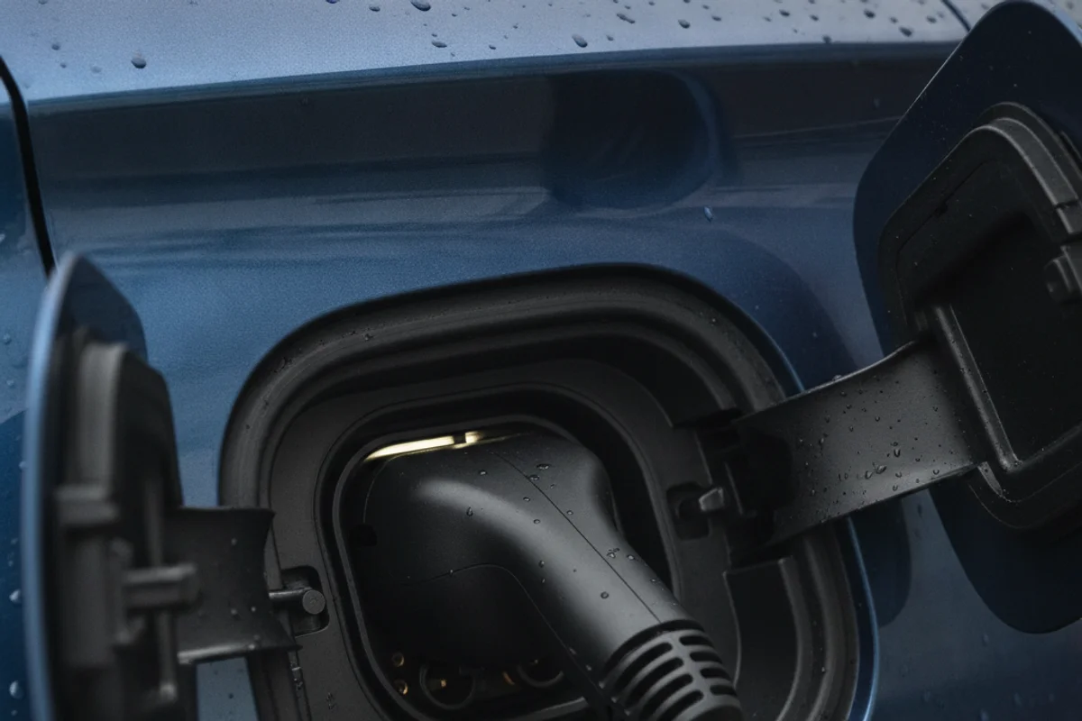 A black charging cable is plugged into a blue electric car, with water droplets visible on the car's surface.