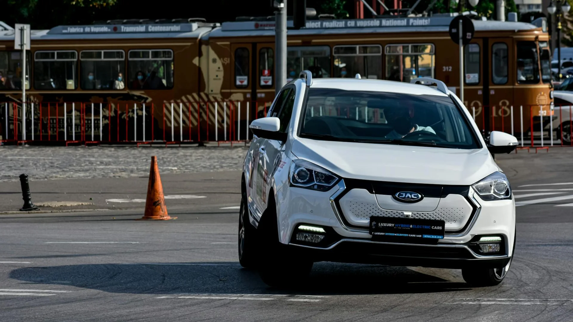 A white electric car with a black grille and illuminated headlights drives on asphalt, with a brown tram in the background.