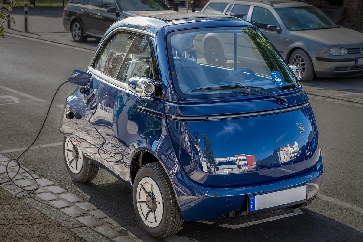 A small, shiny blue electric car is parked on a cobblestone street, charging with a black cable plugged into its side.