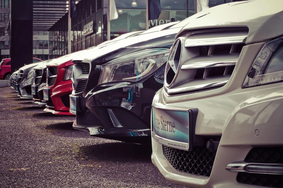 A line of new cars, including white, black, and red models, are parked on asphalt in front of a building.