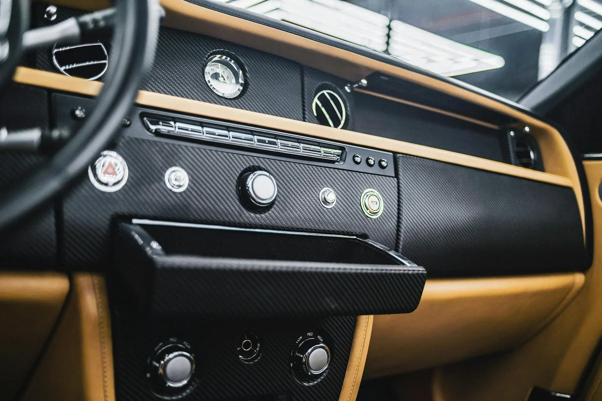 Car dashboard with black carbon fiber and tan leather trim, featuring chrome buttons and a small open compartment.