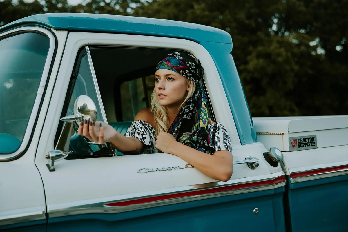A blonde woman in a colorful headscarf leans out the window of a blue and white vintage truck.