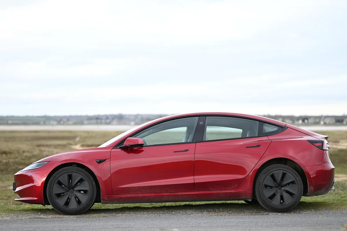 A red Tesla Model 3 is parked on a paved road with a grassy field and cloudy sky in the background.