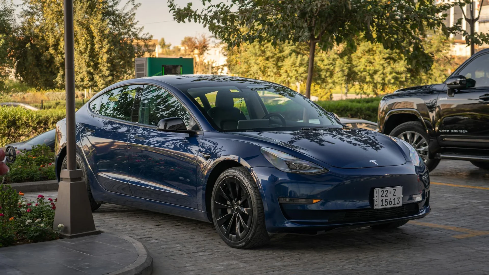 A dark blue Tesla Model 3 is parked on a paved surface, with a black SUV visible behind it.