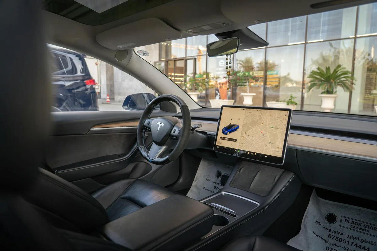 The interior of a Tesla shows a black steering wheel and a large central screen displaying a map and a blue car.
