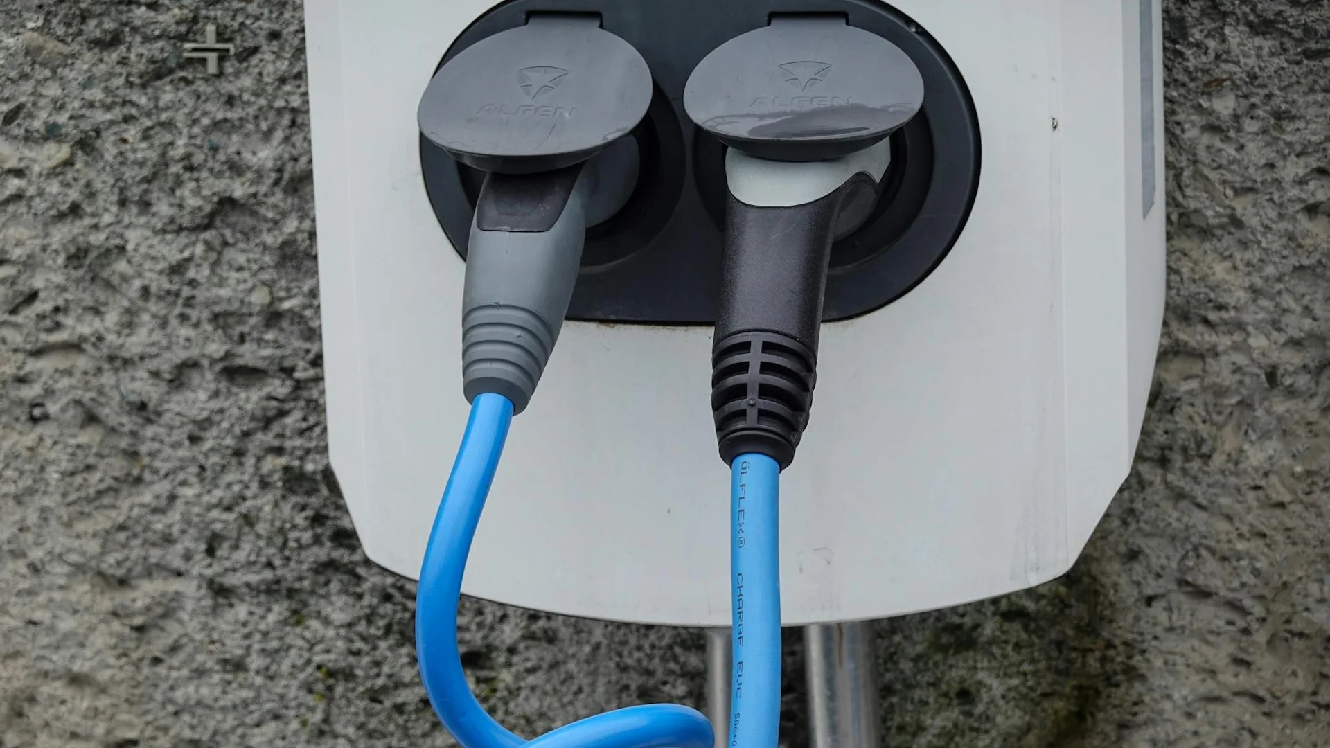 Close-up of an electric vehicle charging station with two cables in sockets against a textured wall.
