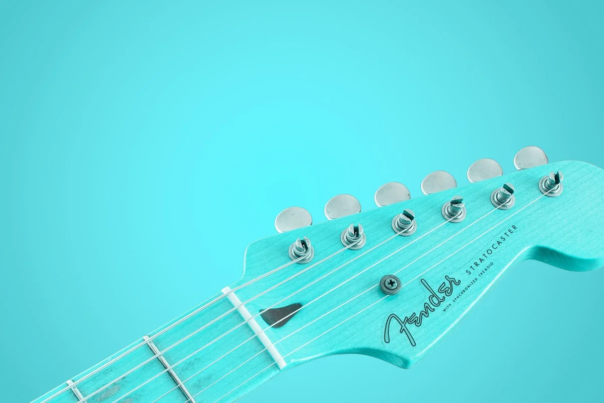 A turquoise Fender Stratocaster guitar headstock with six tuning pegs and strings, angled against a matching background.
