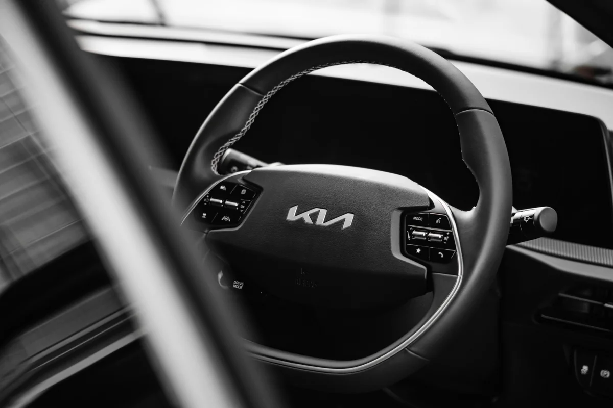 A black and white close-up of a car's steering wheel with a silver Kia logo in the center.