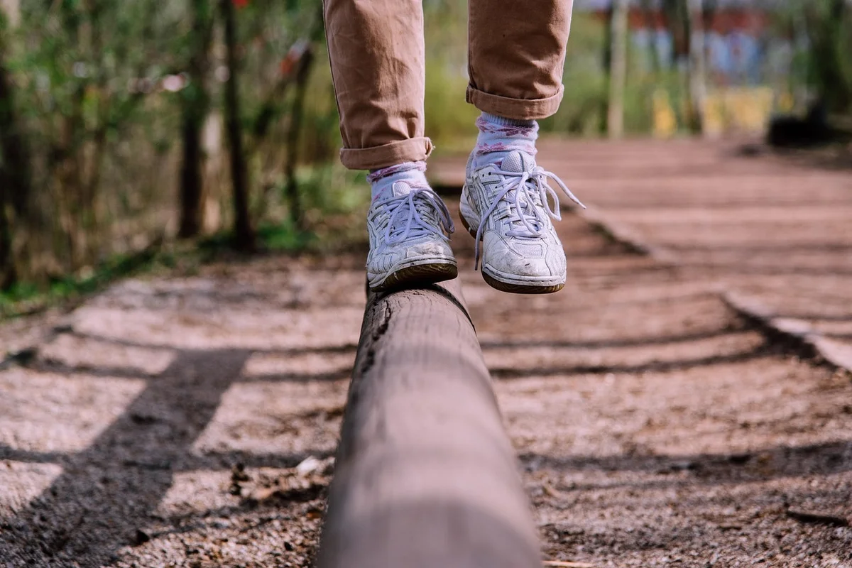 A person in brown pants and white sneakers balances on a dark wooden log, with one foot lifted.