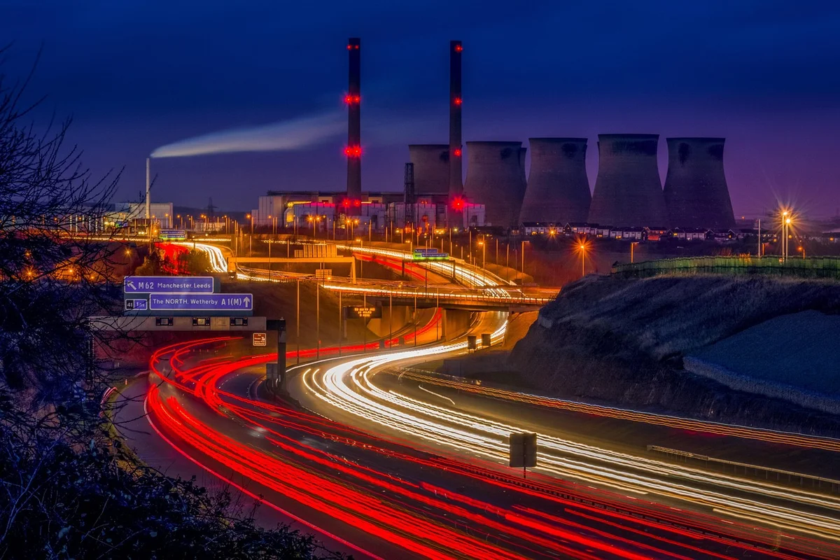 Long exposure of a highway with red and white light trails, leading to a power station with cooling towers at dusk.