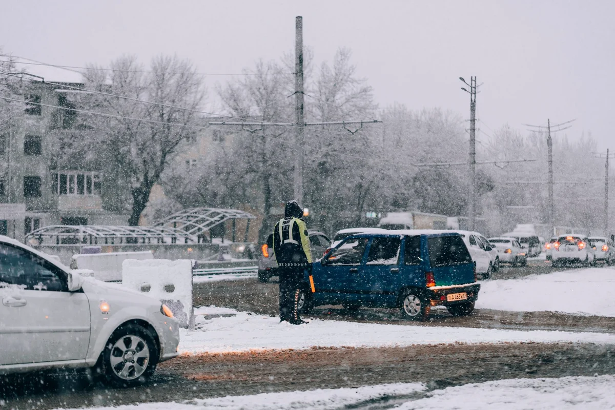 A traffic controller in a yellow vest stands next to a blue car on a snowy road with other cars and buildings.