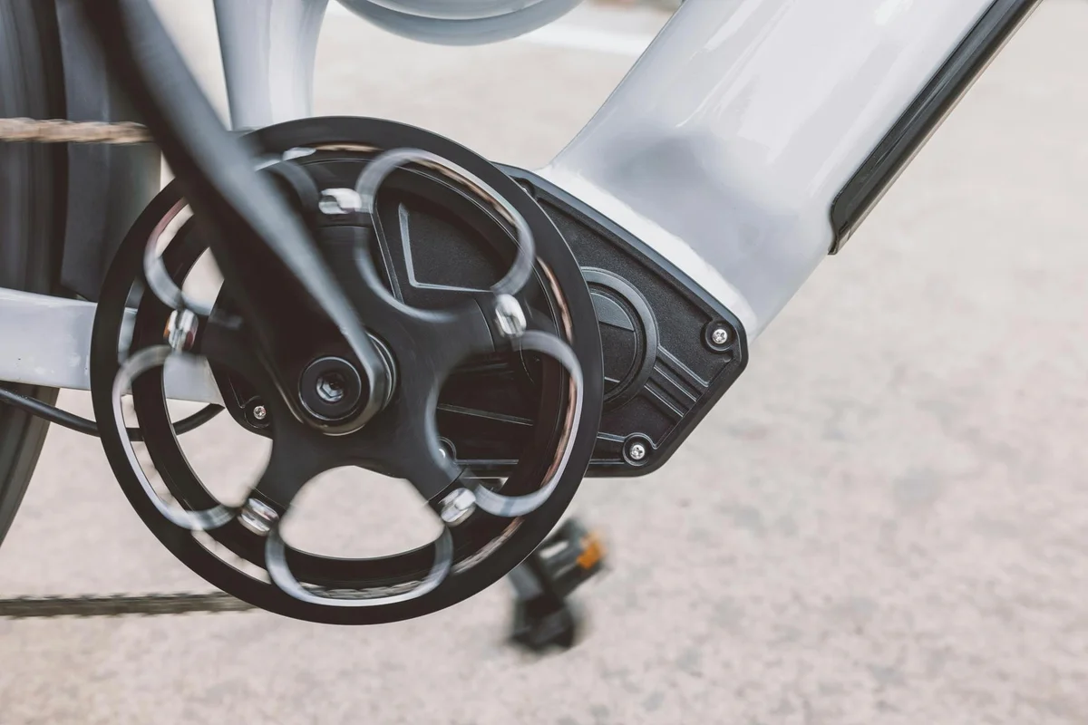 A close-up of a bicycle's black crankset and chain, with a light gray frame and a dark motor housing.