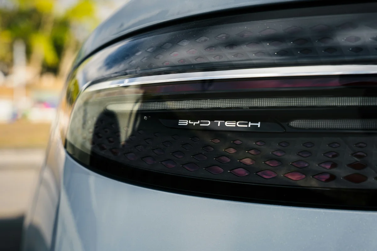 Close-up of a light gray car's dark taillight, featuring a chrome strip and "BYD TECH" text.