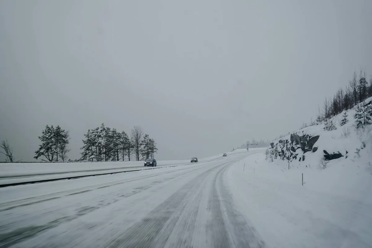 A snow-covered highway with several cars driving, flanked by snow-dusted pine trees on the left and a rocky, snowy hill on the right.