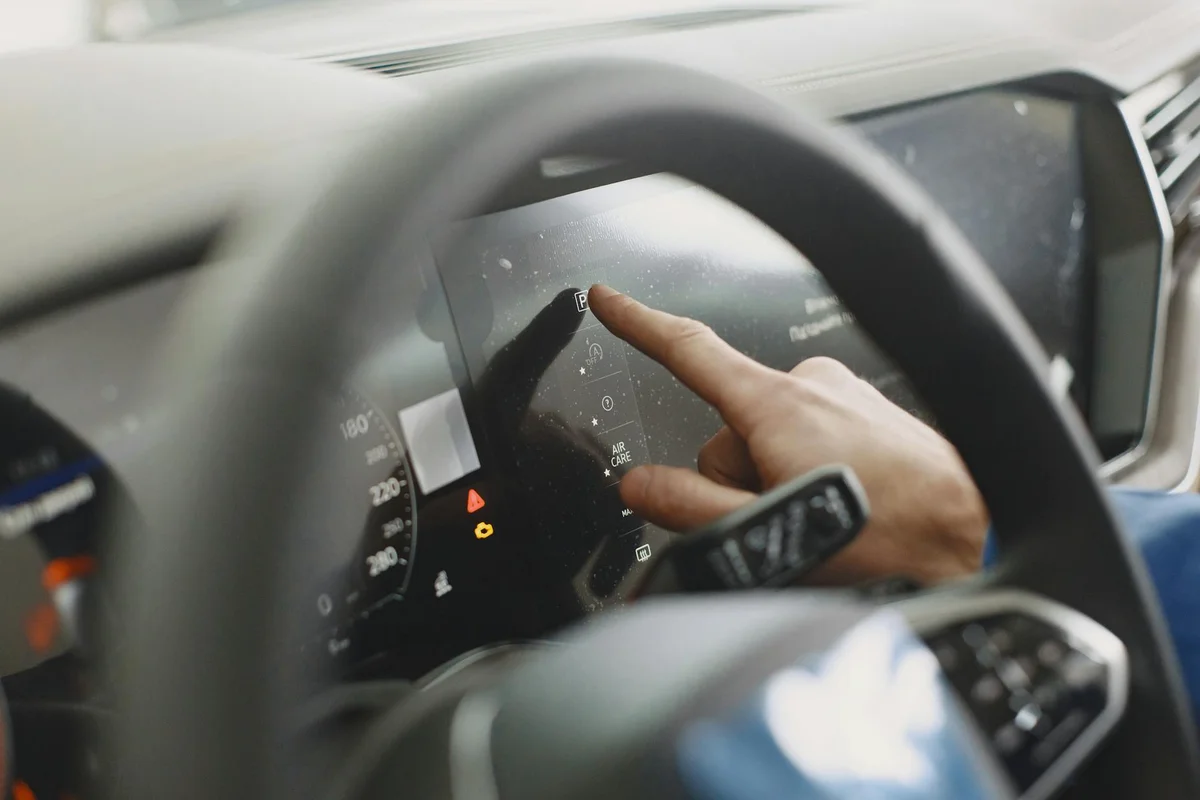 A hand touches a car's dusty black touchscreen display, with a steering wheel in the foreground.