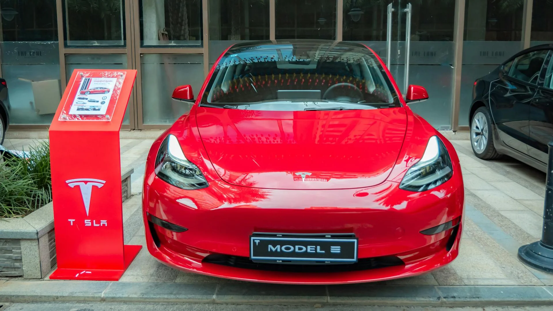 A red Tesla Model 3 is parked in front of a building, with a red Tesla display stand to its left.