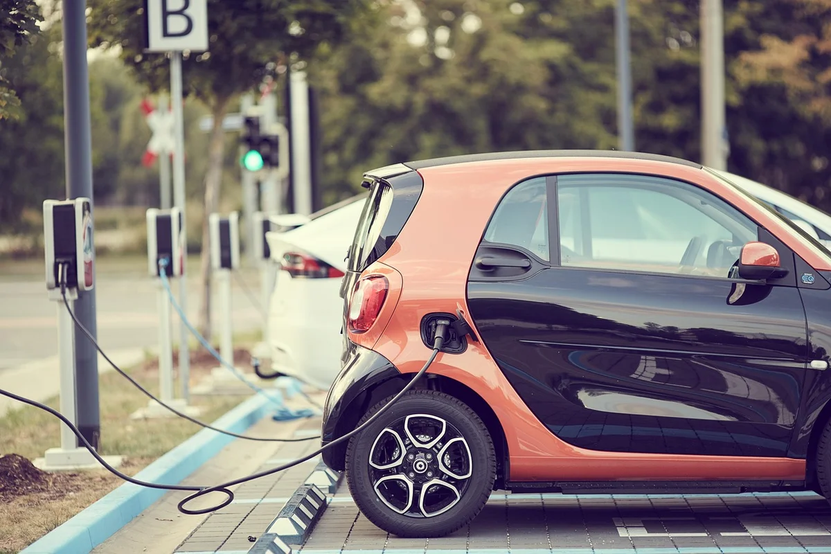 An orange and black electric car is charging at a station, with a white car also charging in the background.