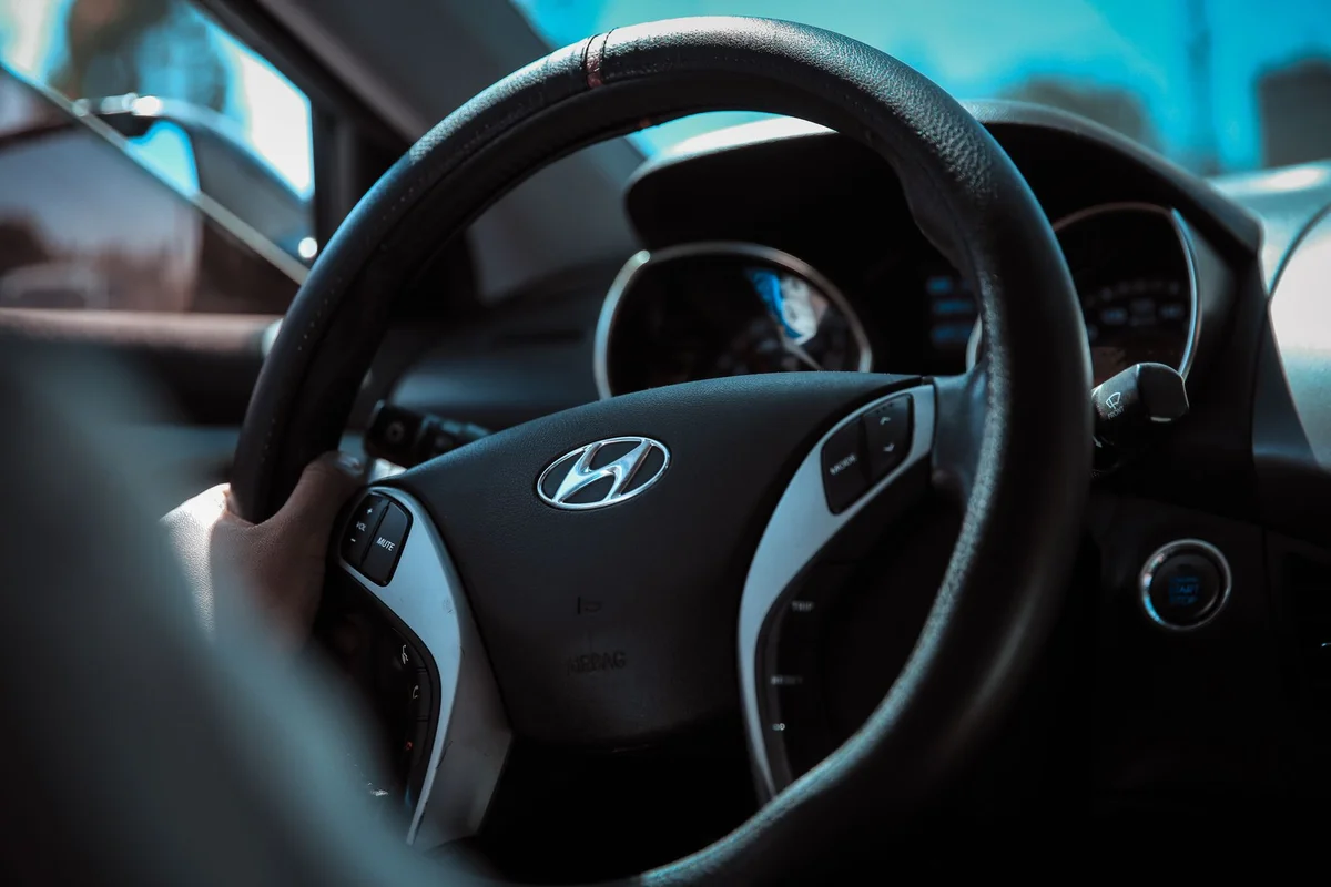 A person's hand grips a black steering wheel with a silver Hyundai logo and control buttons.