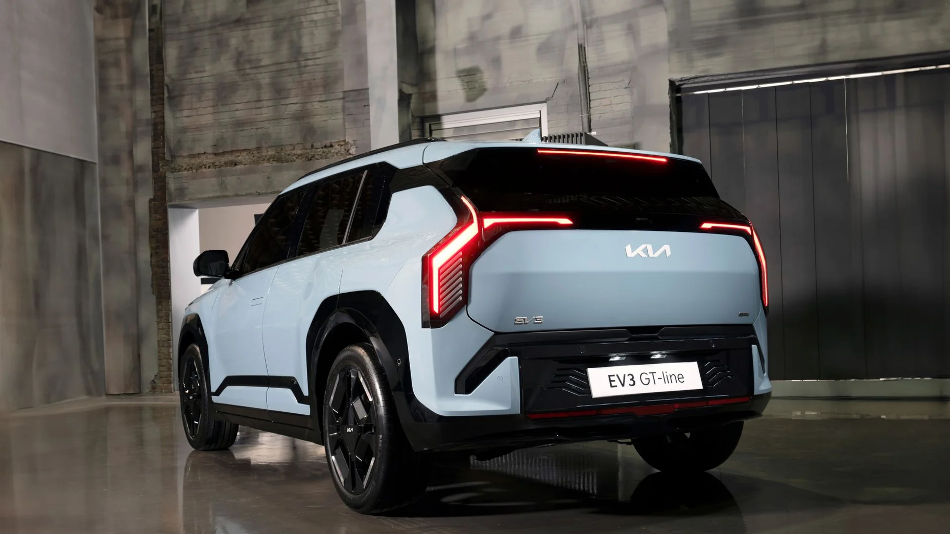 Light blue Kia EV3 GT-line car, seen from the rear, with bright red taillights in a dark garage.