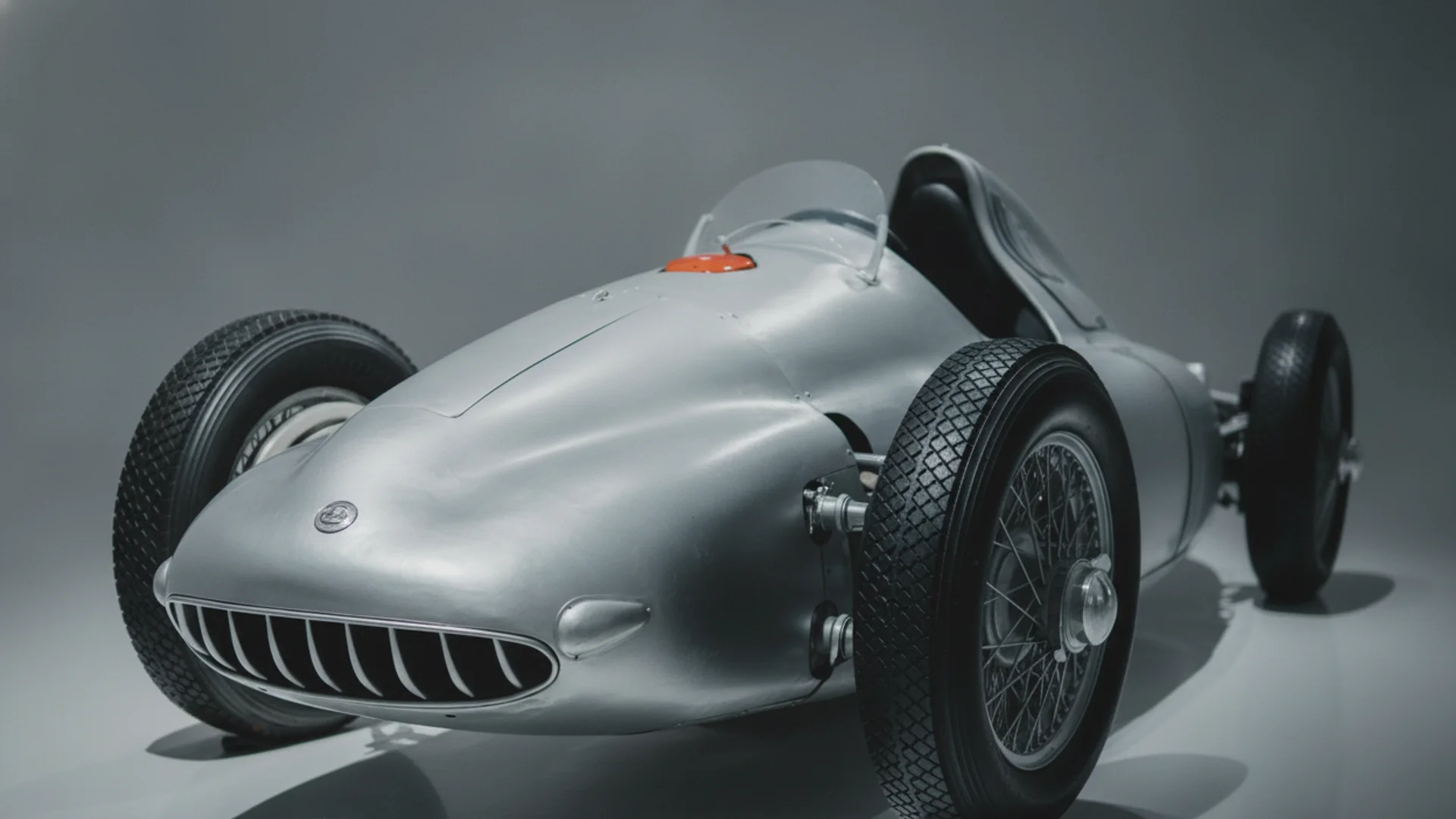 A vintage silver race car with an open cockpit and exposed wheels is angled towards the viewer.