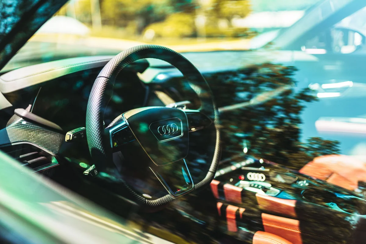 A black Audi steering wheel with the logo is visible through a car window, reflecting trees and sky.
