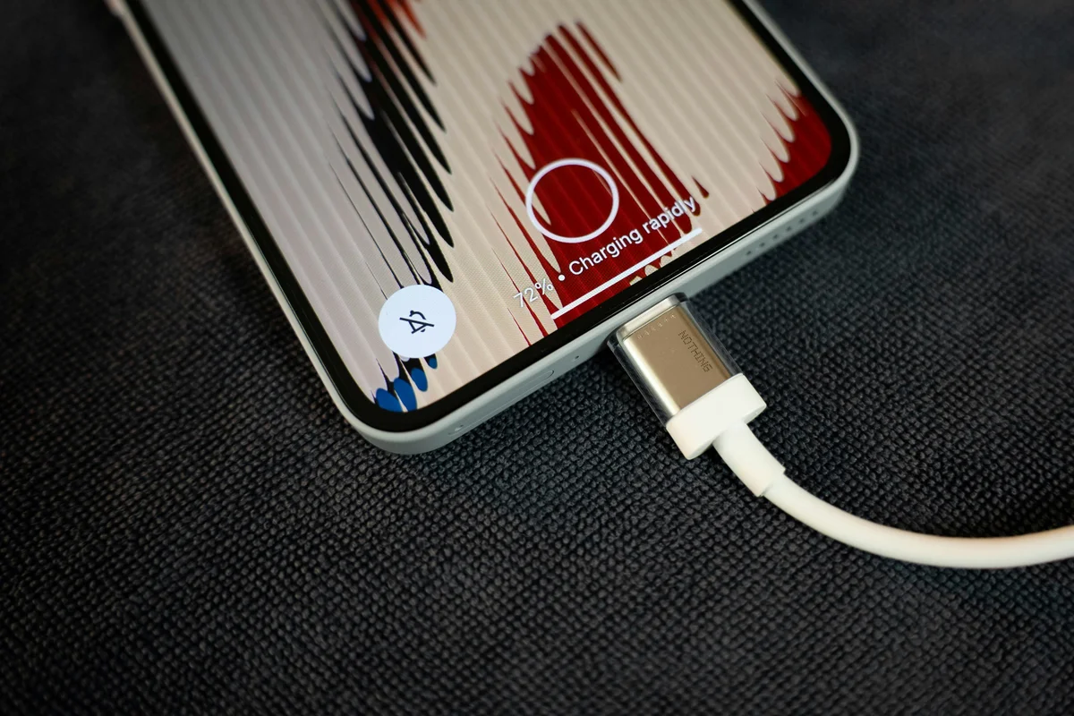 A white smartphone with a red and black striped background is plugged into a white "NOTHING" brand charger.