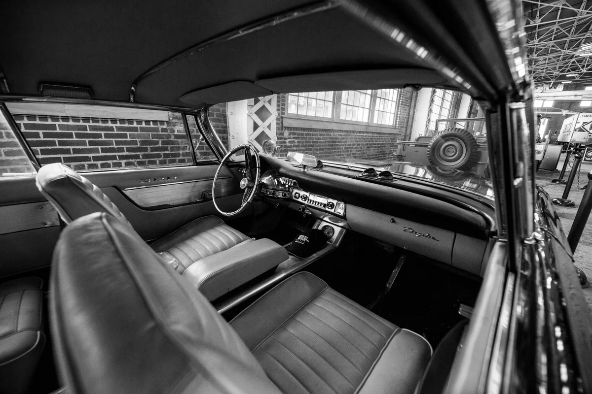 The black and white interior of a vintage car shows the steering wheel on the right and two rows of seats.