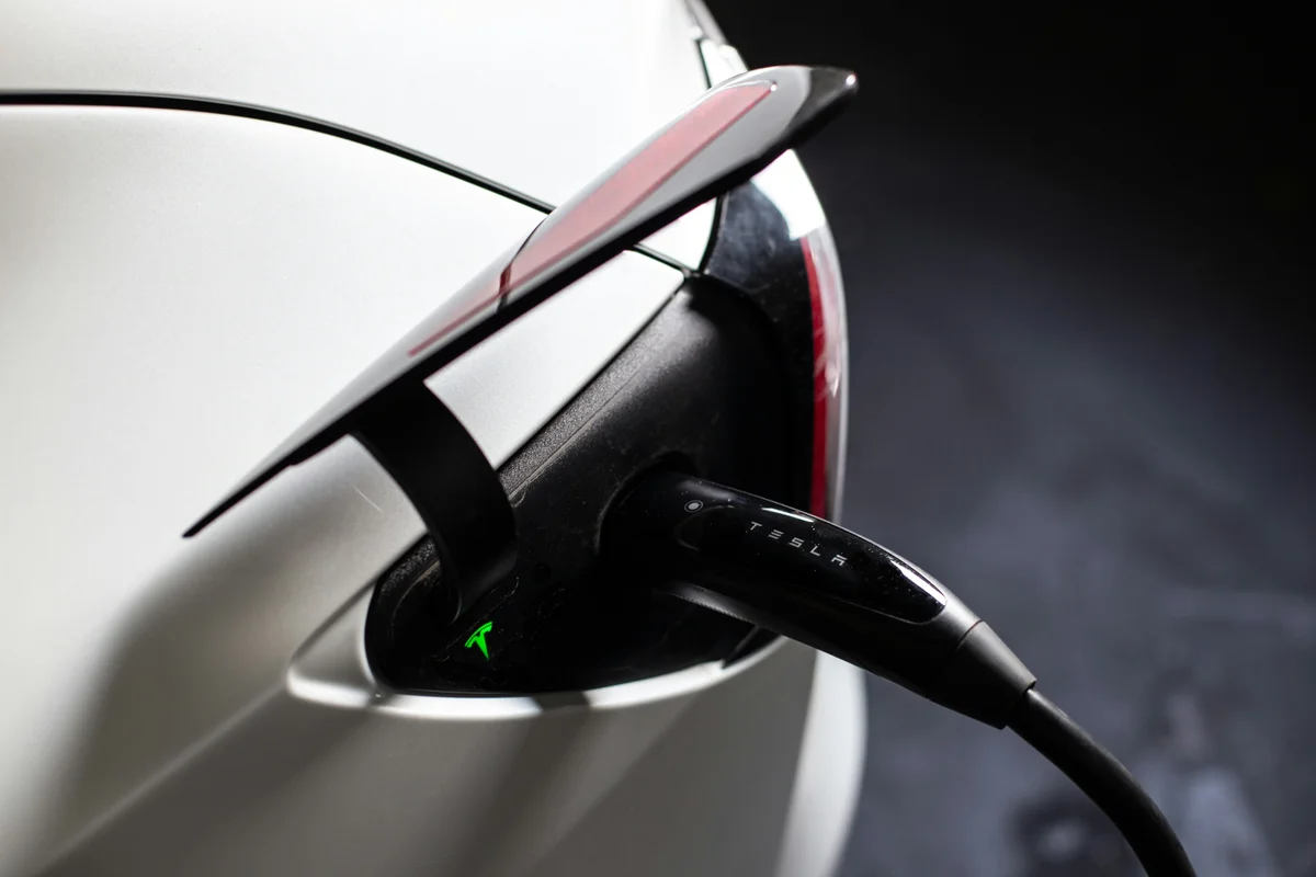 A black Tesla charging cable is plugged into the back of a white Tesla car, with a green light on the port.