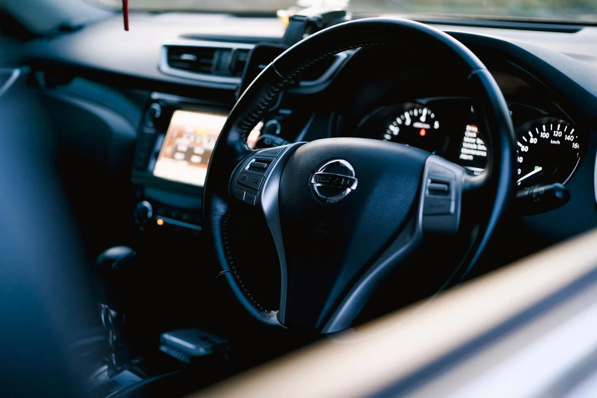 A black Nissan steering wheel with a silver logo and control buttons is in the foreground of a car interior.