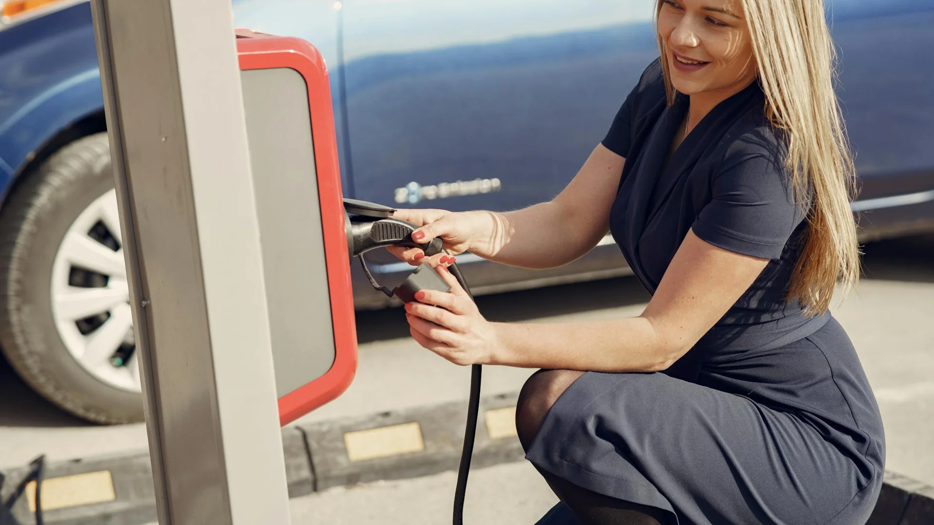 A smiling woman in a dark dress crouches, plugging a black charging cable into a red and gray charging station.