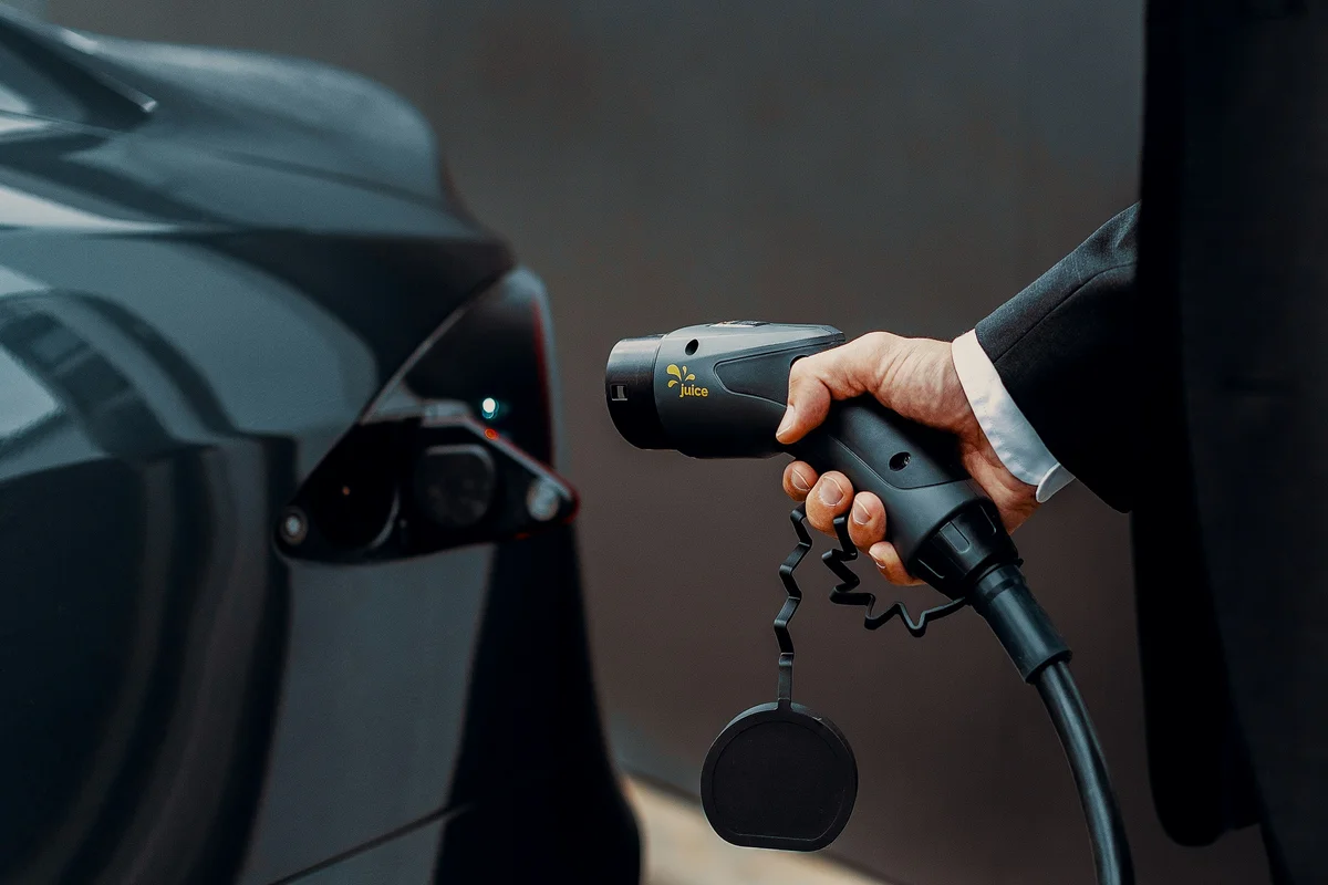 A person's hand in a suit jacket holds a black Juice brand EV charger, aiming it at a dark grey car's charging port.
