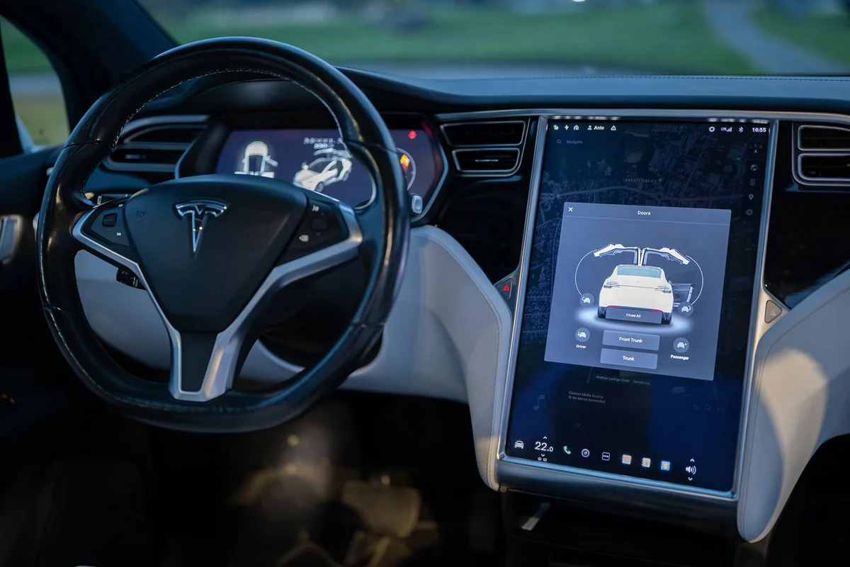 A Tesla car interior shows a black steering wheel and a large touchscreen displaying car controls.