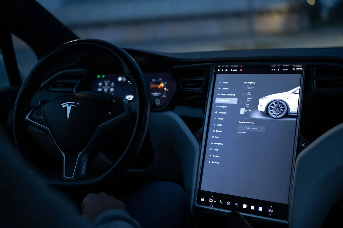 A Tesla interior shows a steering wheel on the left and a large touchscreen displaying vehicle settings on the right.