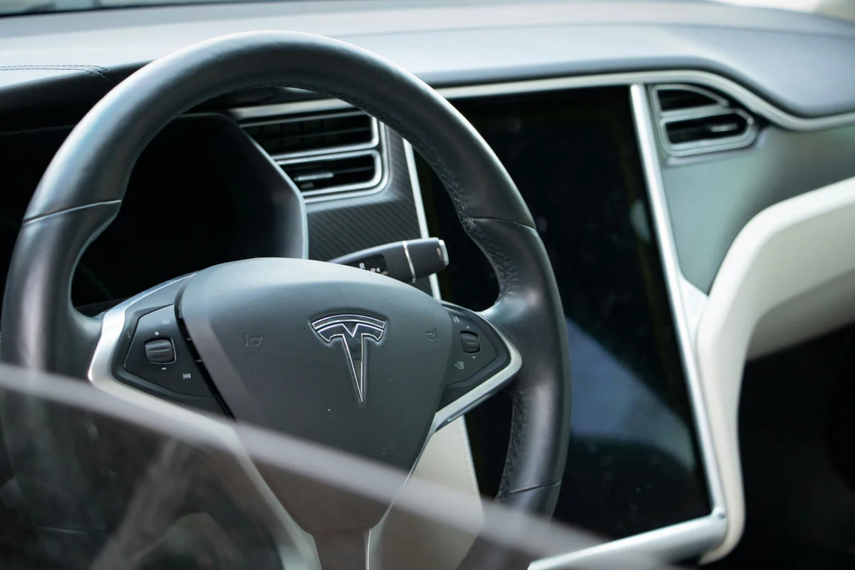 A black leather steering wheel with a silver Tesla logo in the center, and a large dark screen to its right.