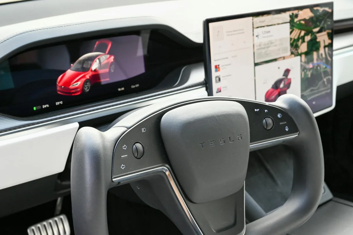 A black Tesla steering wheel with a screen showing a red car with open doors on the left and a map on the right.
