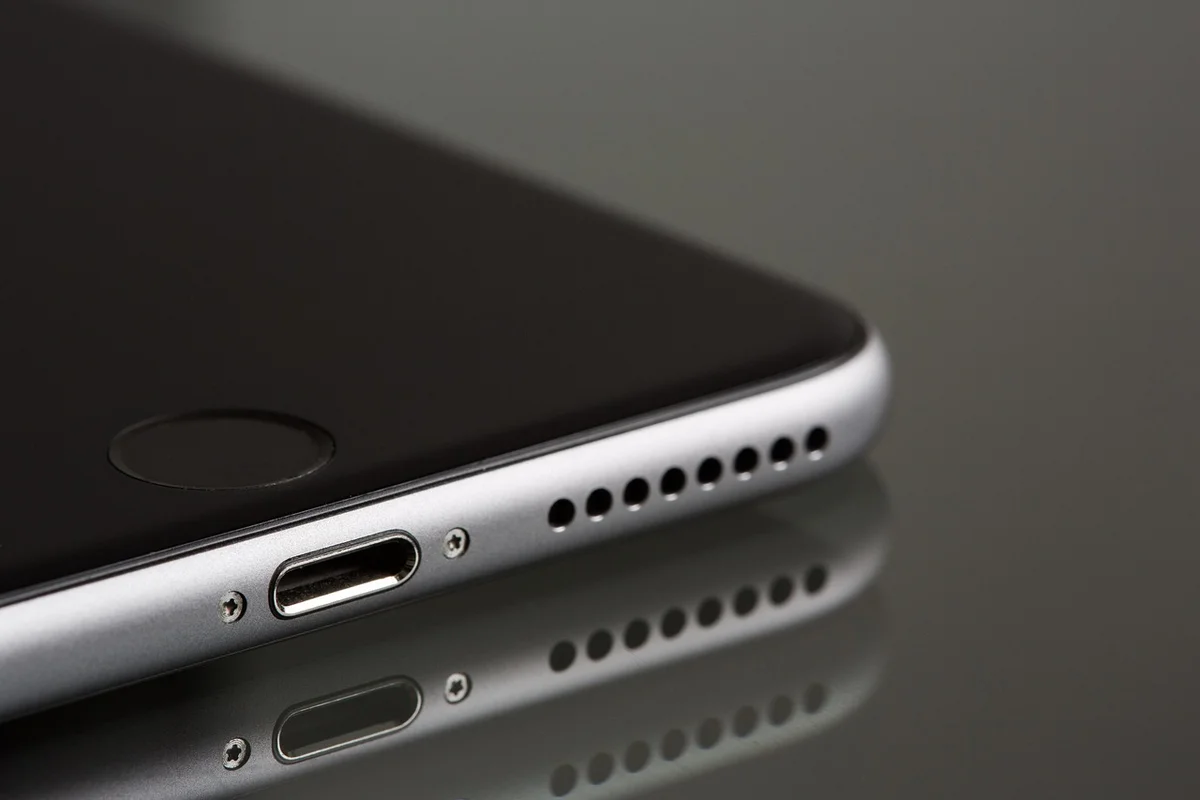 A silver smartphone with a black screen is angled on a reflective surface, showing its charging port and speaker holes.