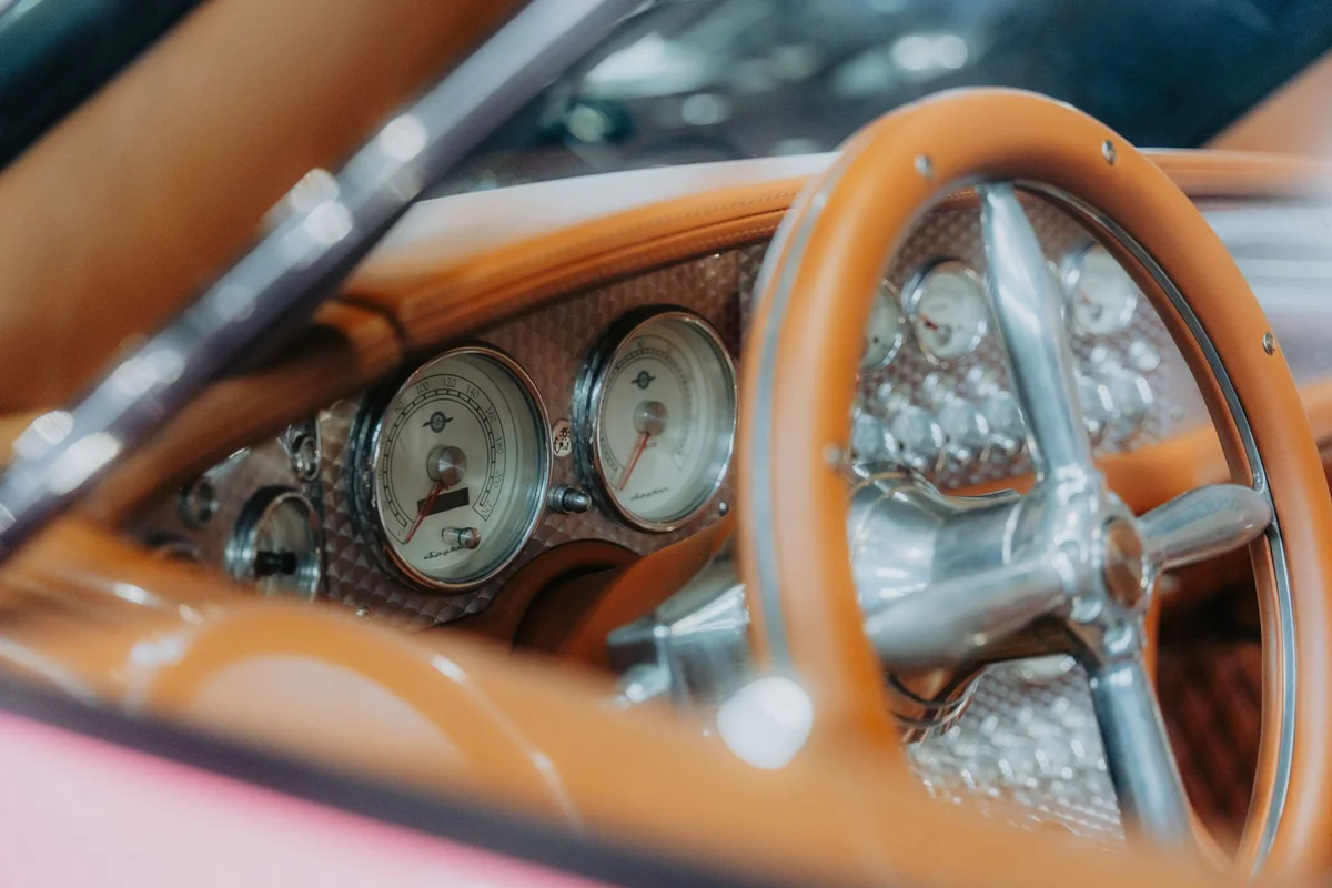 A close-up of a car's dashboard with a tan leather steering wheel and two round gauges on a textured silver panel.