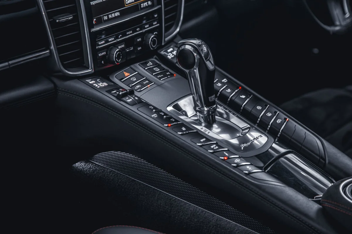 A car's dark interior with a silver gear shifter in the center console, surrounded by black buttons with red lights.