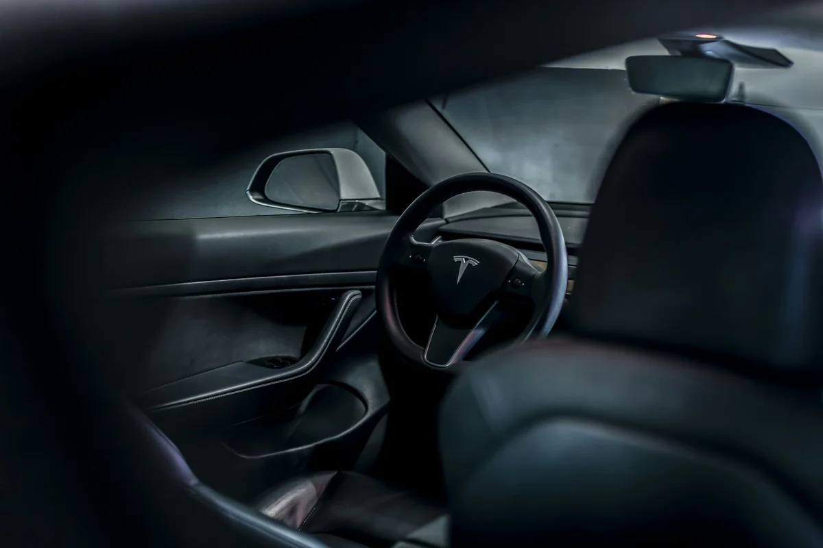 The dark interior of a Tesla car with a steering wheel and side mirror visible.