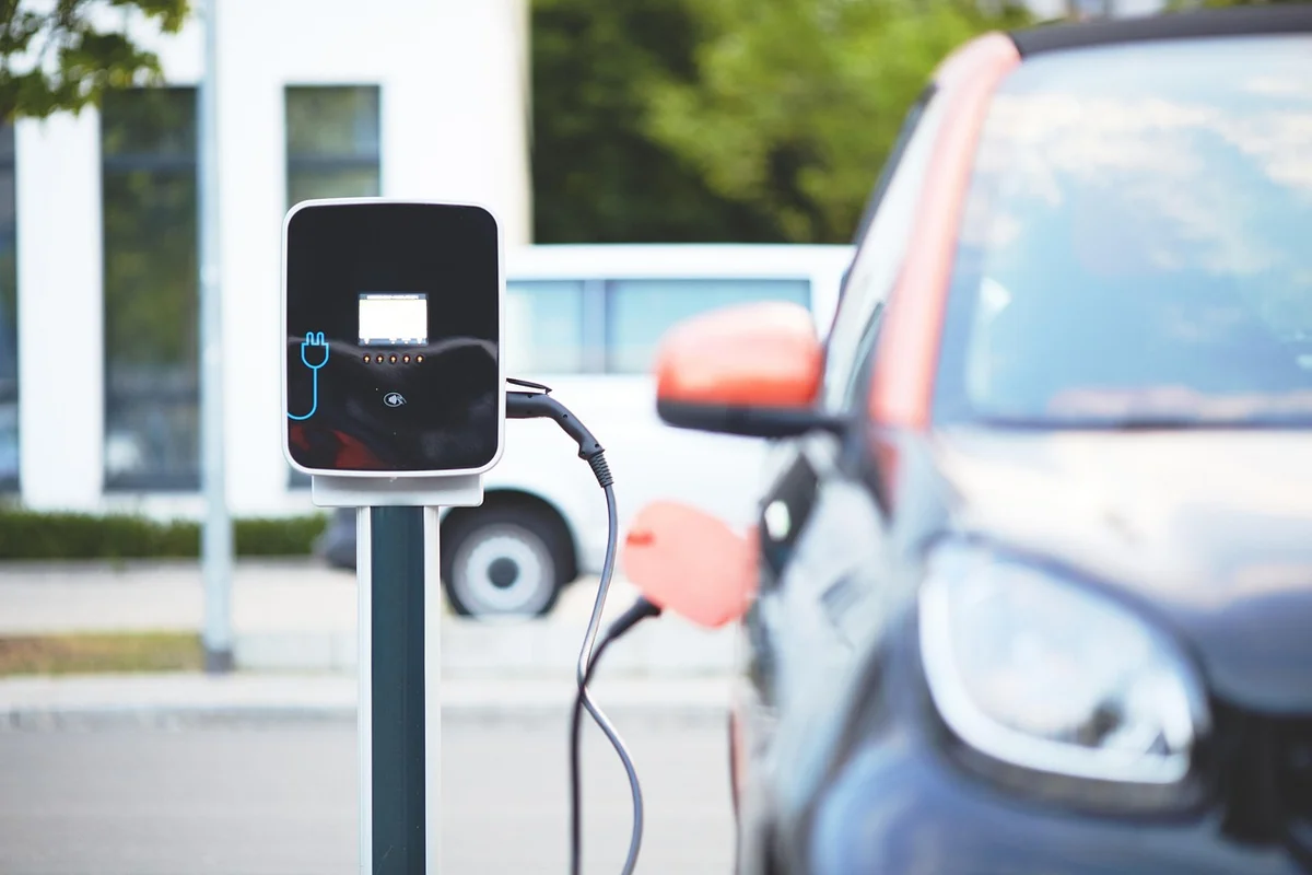 An electric car with an orange side mirror is plugged into a black charging station on the left.