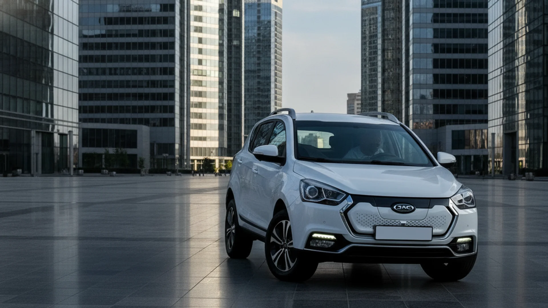 A white electric SUV is parked on a paved plaza with tall glass buildings in the background.