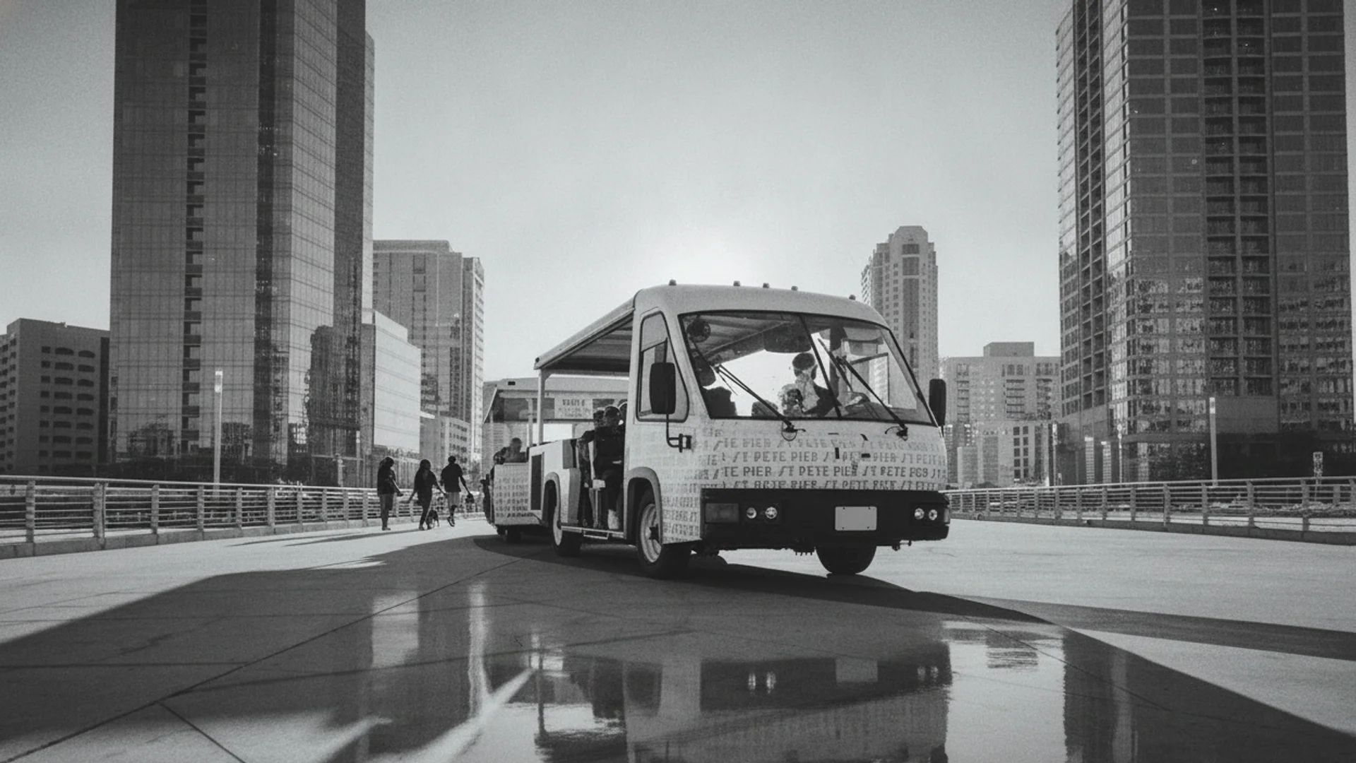 A black and white image of a white open-air shuttle bus driving right on a paved road with tall buildings.